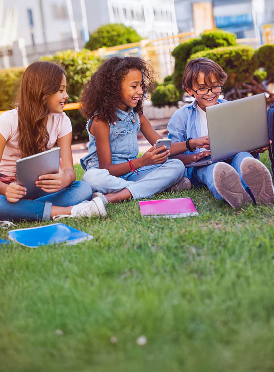 Happy little school children elementary middle school pupils schoolmates with digital gadgets laptop tablet smart phone backpacks notebooks sitting at the grass park outdoors doing homework