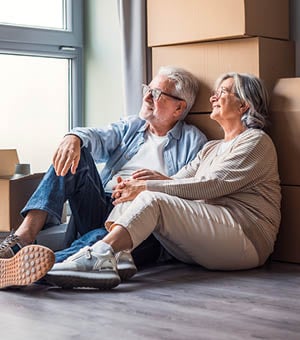Senior couple sit on the floor beside moving boxes, smiling and holding hands as they unpack and settle into their new home, enjoying natural light, calm atmosphere and shared memories.