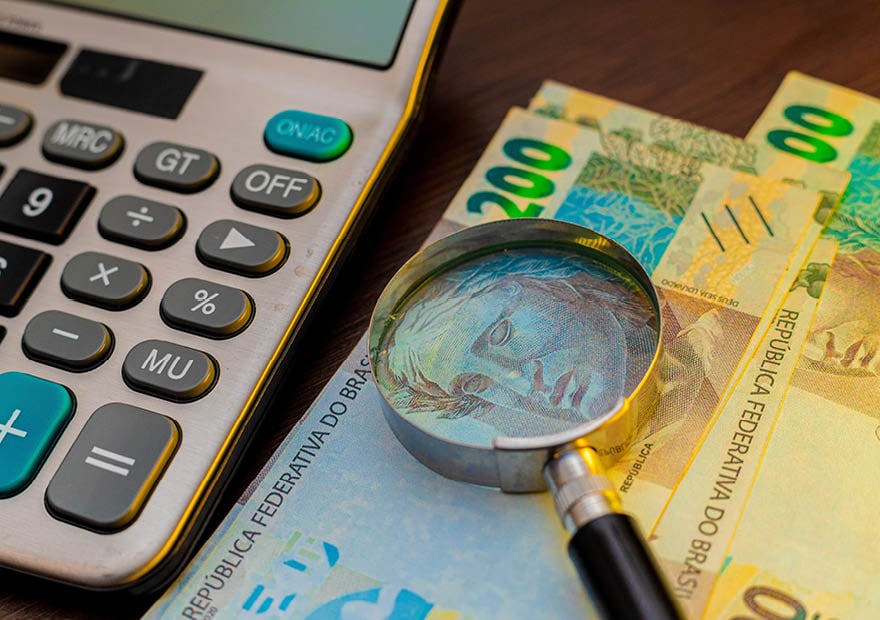 Image of a magnifying glass rests on Brazilian 200 Reals banknotes next to a calculator, suggesting financial analysis, costs, or an inspection of money.