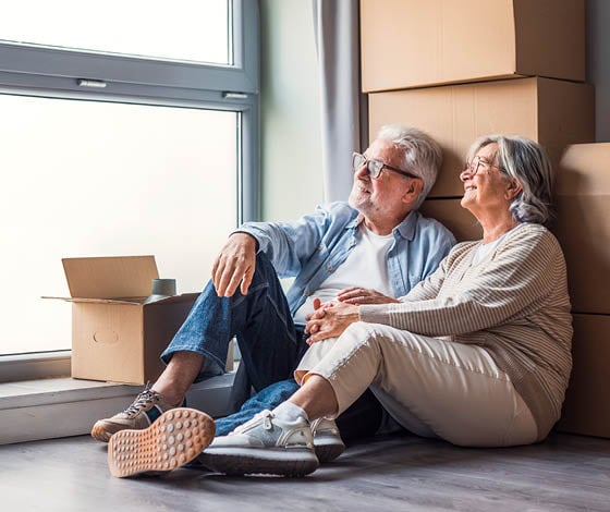 Senior couple sit on the floor beside moving boxes, smiling and holding hands as they unpack and settle into their new home, enjoying natural light, calm atmosphere and shared memories.